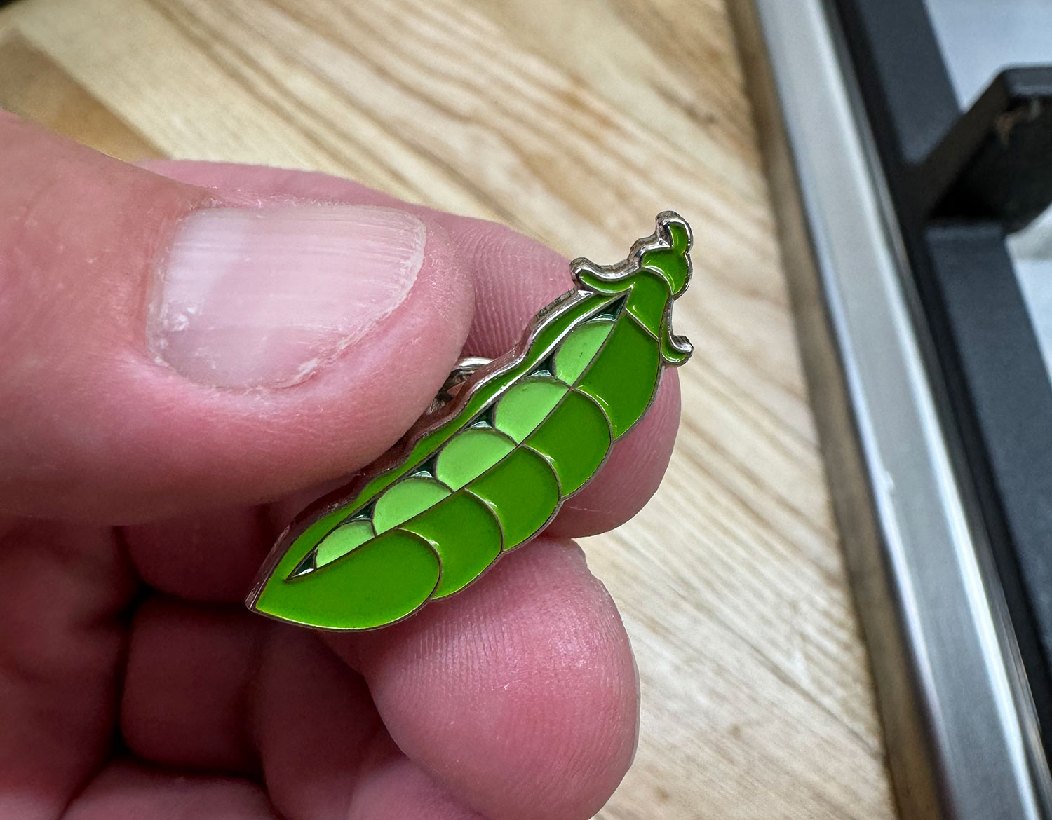Pea emoji enamel pin badge held by a thumb and two fingers. There is a wood grain desk top visible in the background