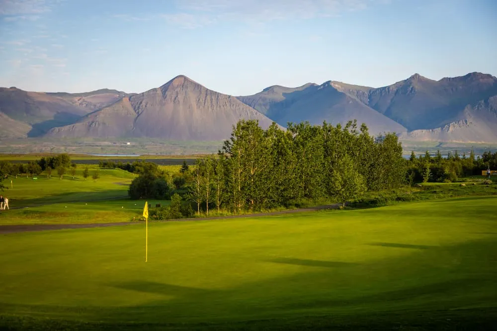 A lush green golf course with a yellow flag in the foreground, a row of tall trees, and a backdrop of large, rocky mountains under a blue sky.