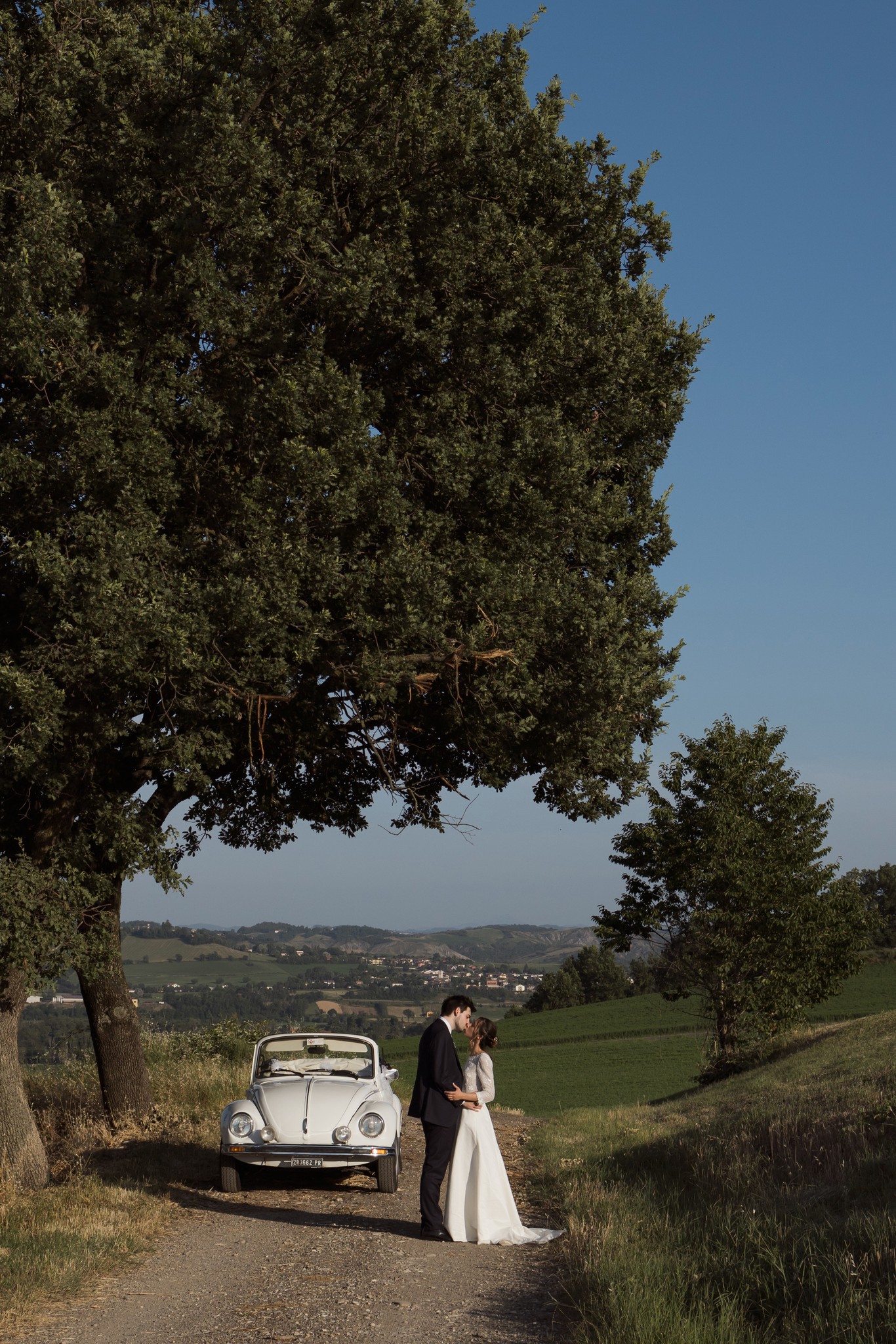 Sposi al fianco di un Maggiolino bianco tra i sampietrini, paesaggio collinare toscano di Villa Peroni Riva sullo sfondo