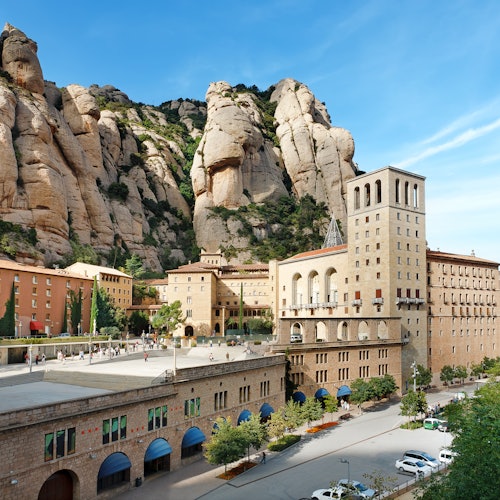 Historic buildings and a plaza at the base of a rocky mountain, with a clear blue sky and parked cars in the foreground.