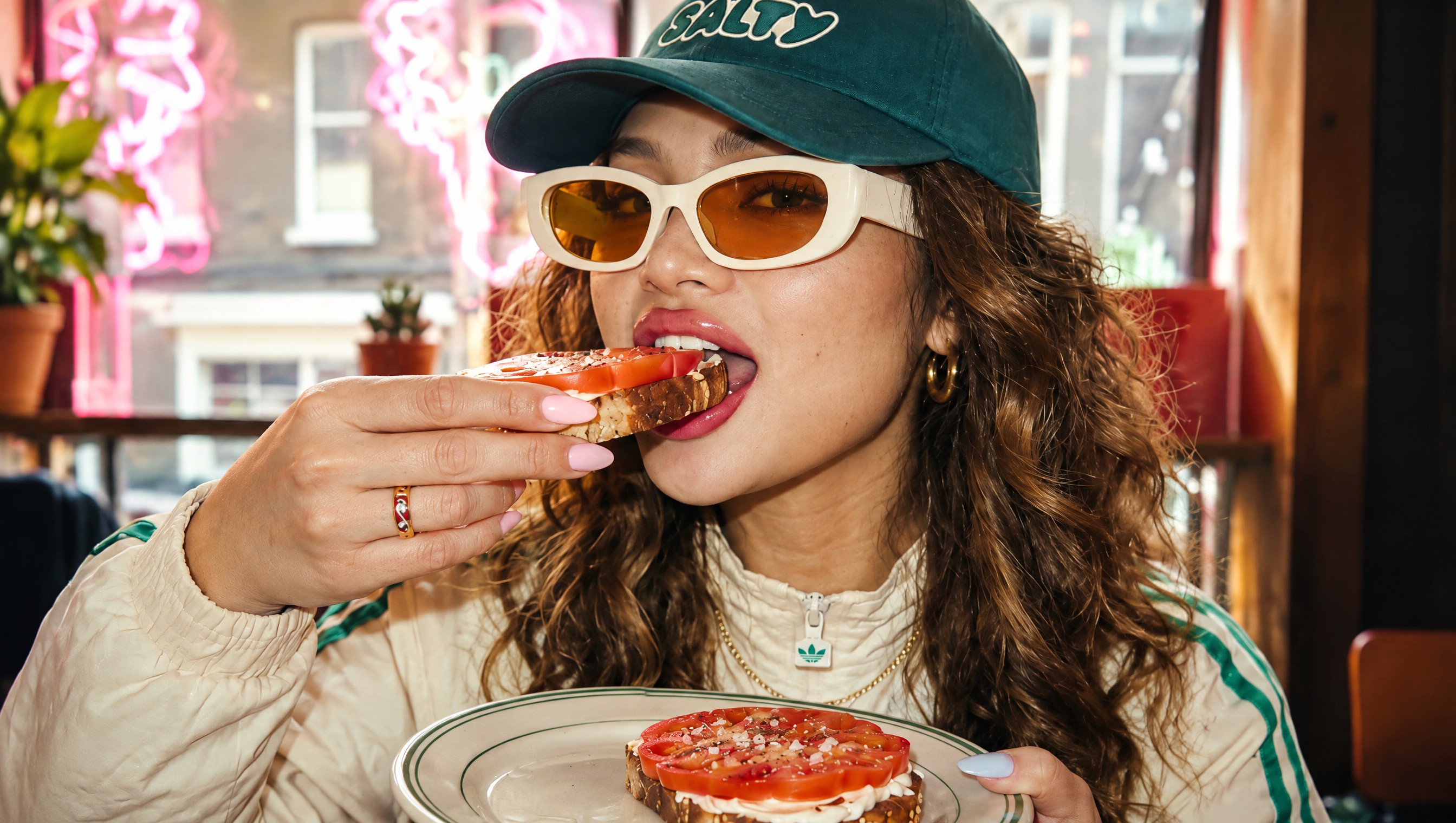 A woman in a Salty cap and white sunglasses biting a tomato bagel in a diner