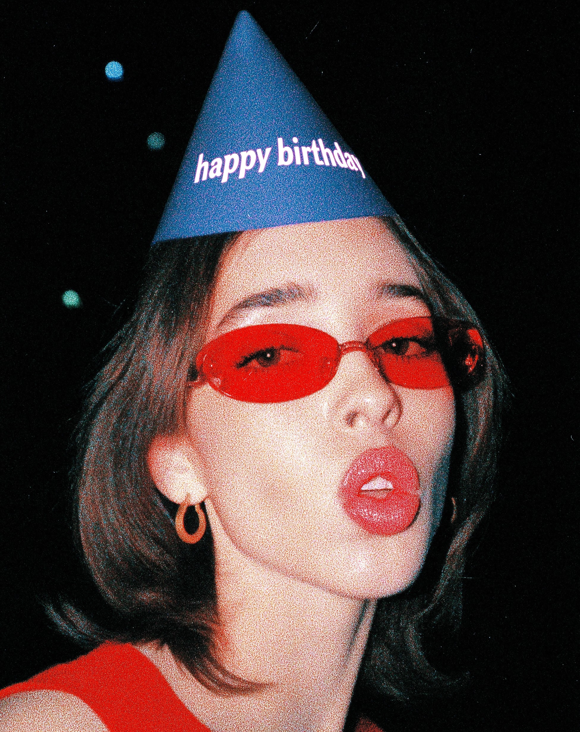 A grainy close-up photo of a woman wearing red oval sunglasses, a blue birthday cone hat, hoop earrings, and a red top, posing against a dark background.