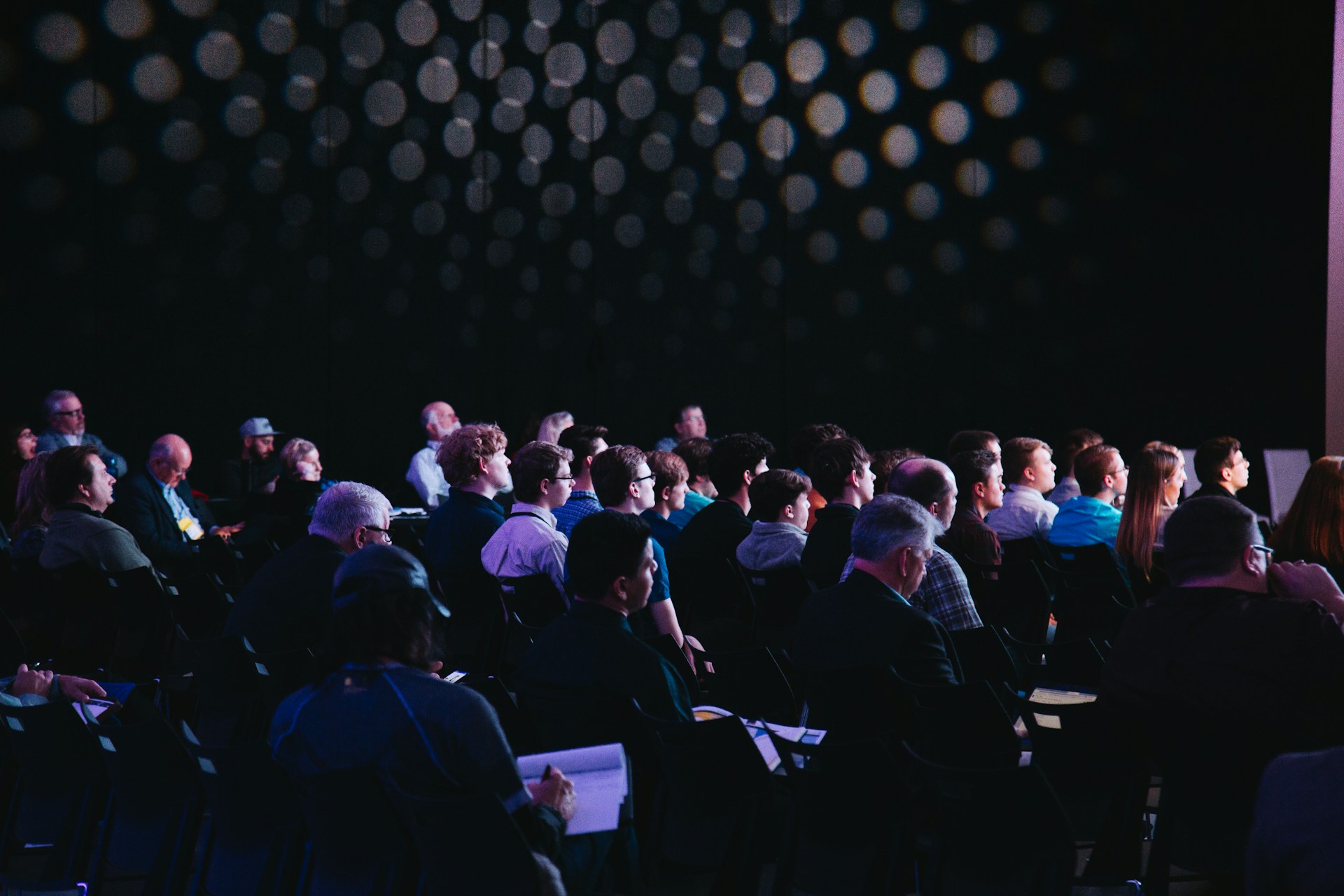A crowd sat in a dark conference hall listening to a presentation 