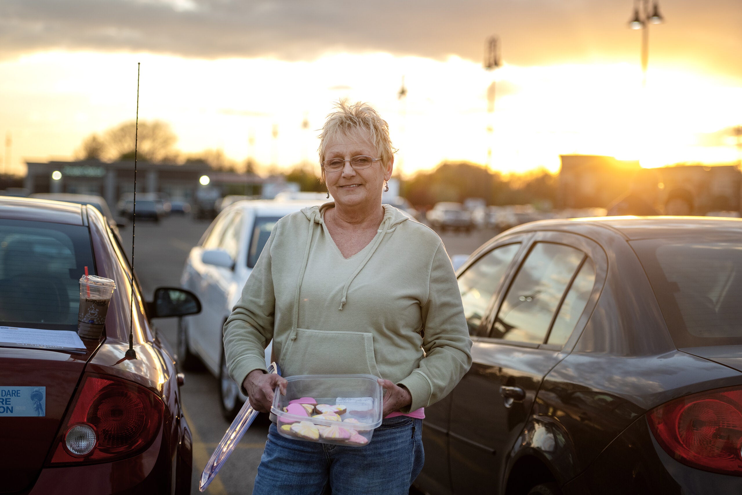 An older woman stands in a parking lot at sunset, holding a container of decorated cookies between two parked cars.