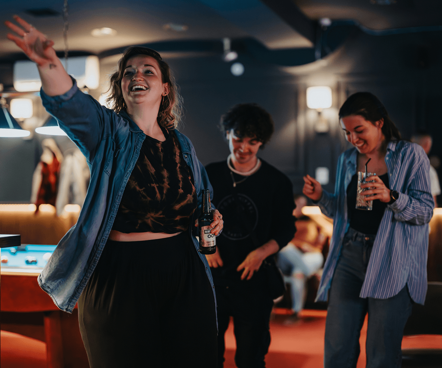 A woman laughs and gestures excitedly while holding a beer, socializing with friends in a dimly lit pool hall.