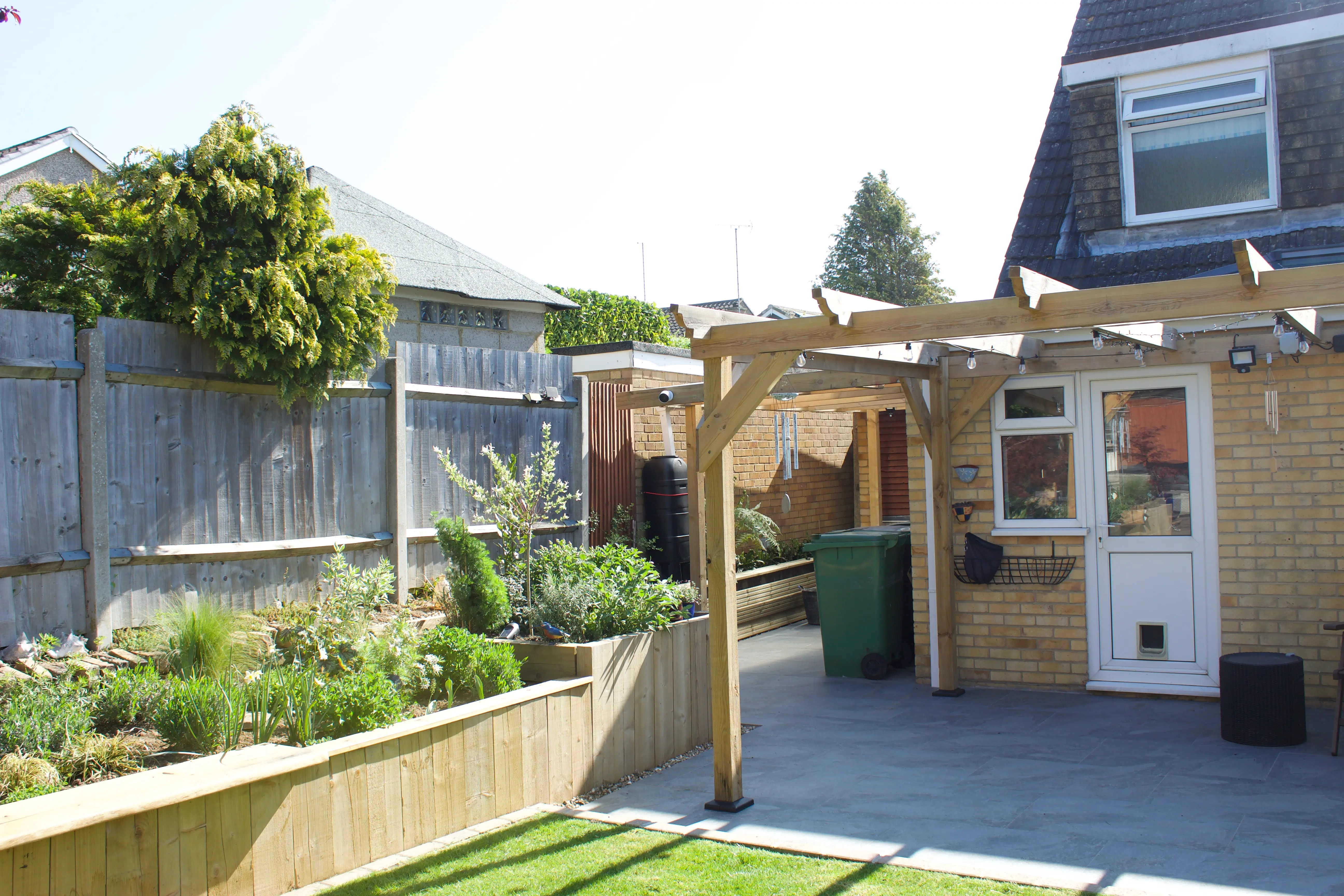 A sunny backyard featuring a wooden pergola, garden beds, and a house in the background.