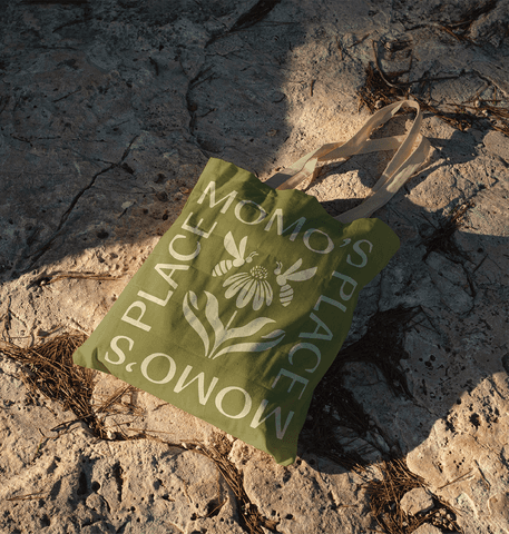 Green canvas tote bag with Momo’s Place logo and botanical bee illustration on rocky ground
