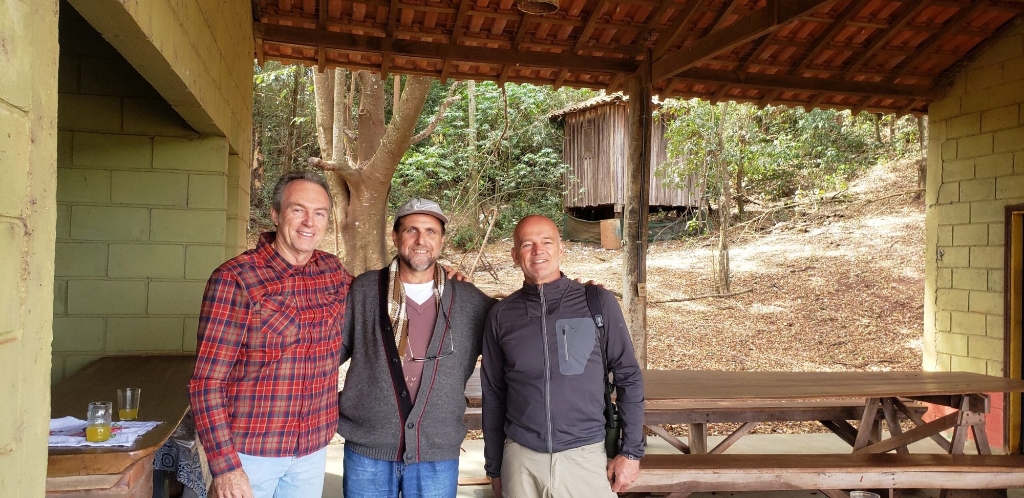 Três homens abraçados posando sob varanda coberta com mesas de madeira e árvores ao fundo