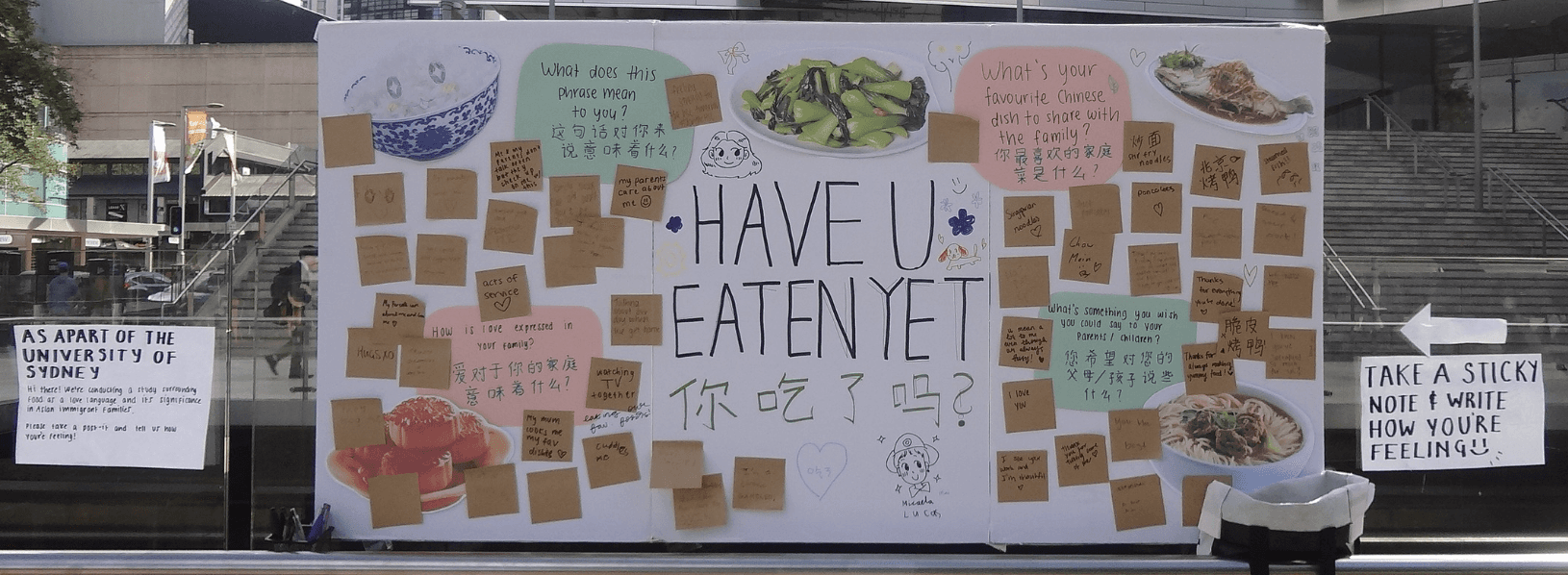 A large public installation board titled “Have U Eaten Yet?” displayed in a city plaza. The board is covered with handwritten sticky notes in English and Chinese, alongside food images and prompt questions like “What’s your favourite Chinese dish to share with family?” and “What does this phrase mean to you?” A sign on the right invites passersby to “Take a sticky note & write how you’re feeling :)”.