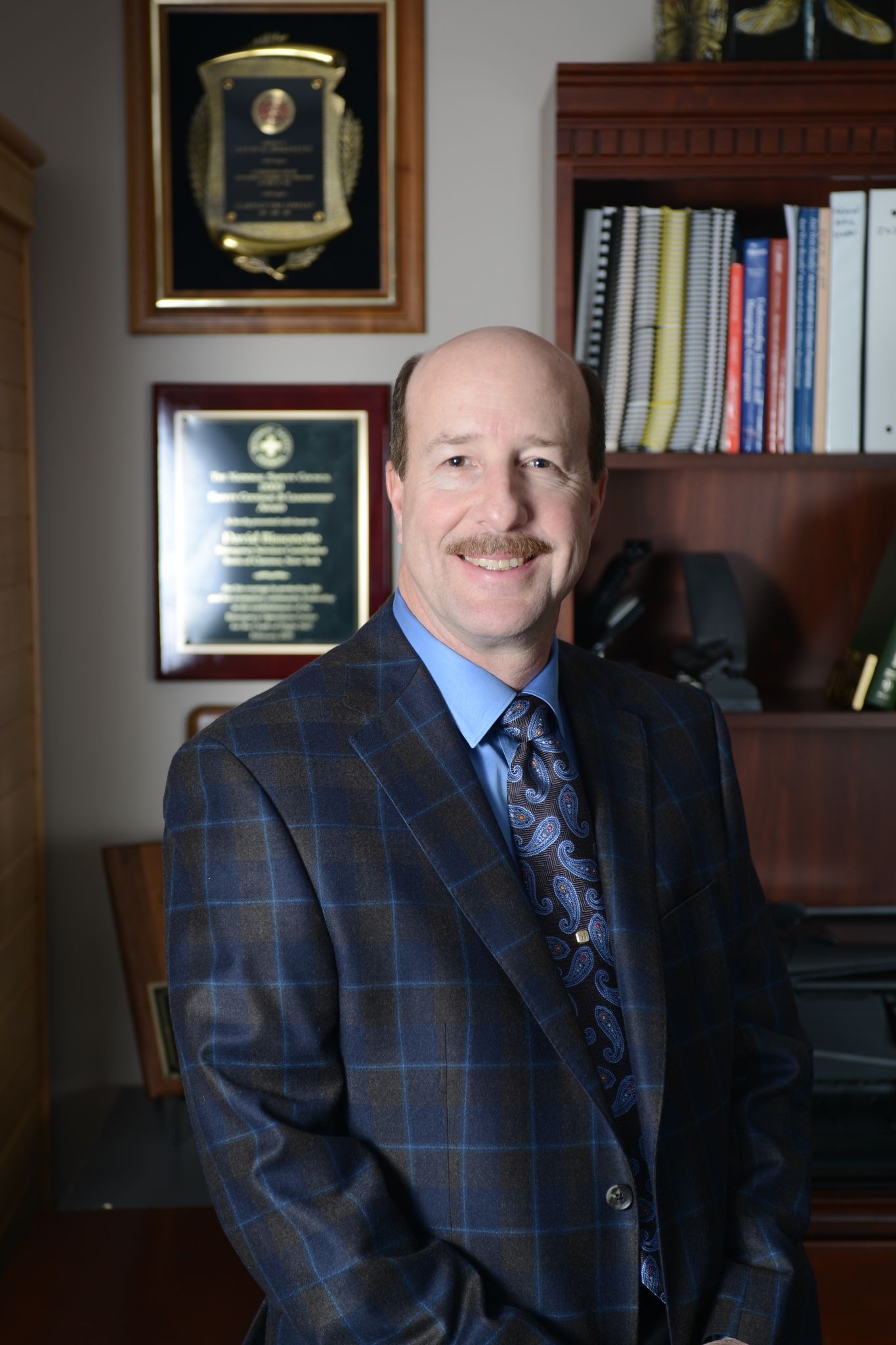 David Bissonette standing in office beside bookshelf and awards, wearing blue plaid suit and paisley tie.