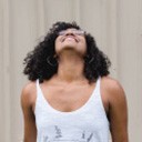 A young woman with curly hair looks up with a joyful expression. She's wearing a white tank top and glasses.
