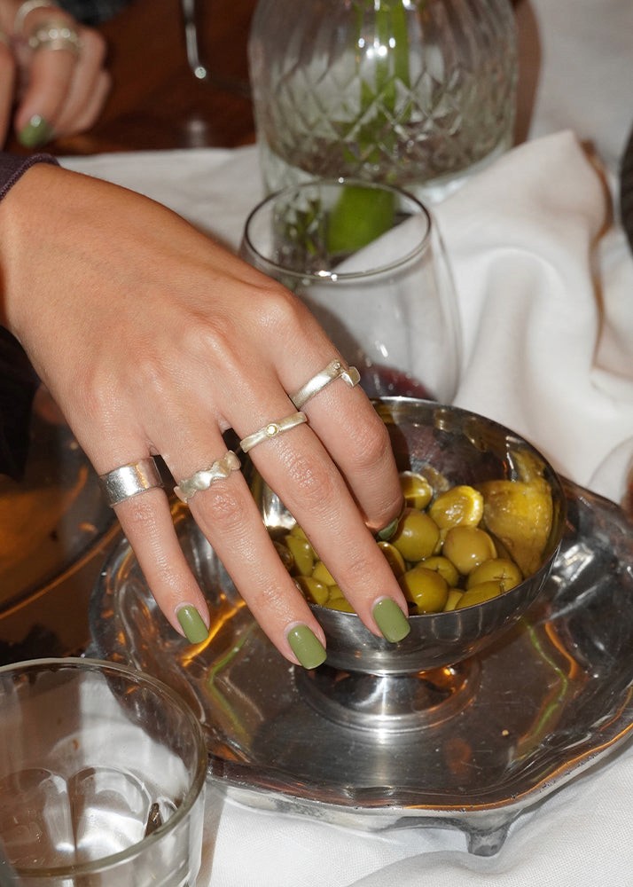 Hand with green nail polish and multiple rings on a silver dish with olives, surrounded by glasses on a table.