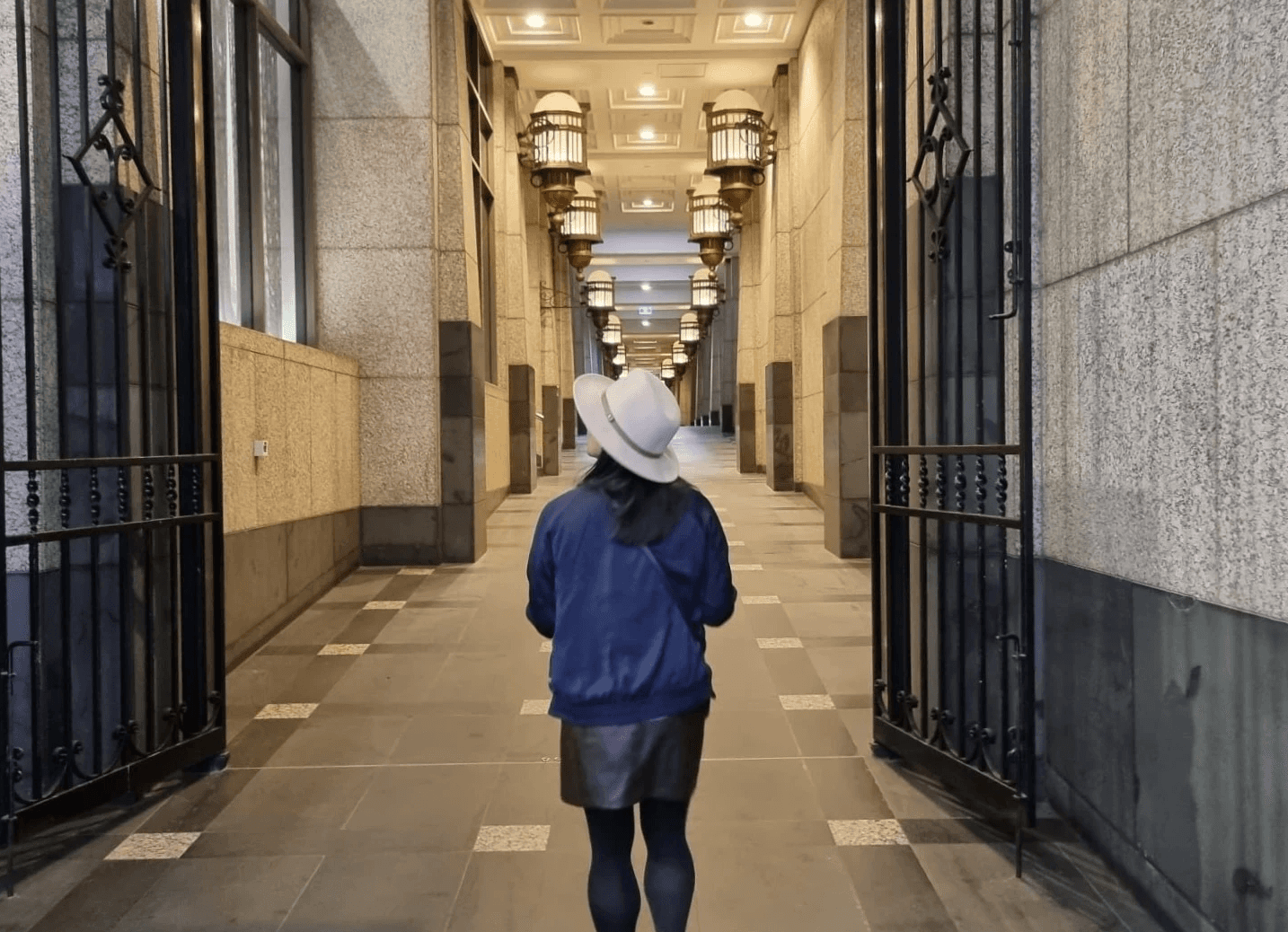 Ann Luong in Melbourne city wearing a beige fedora, navy bomber jacket and brown leather skirt. Looking up at the lights in a laneway