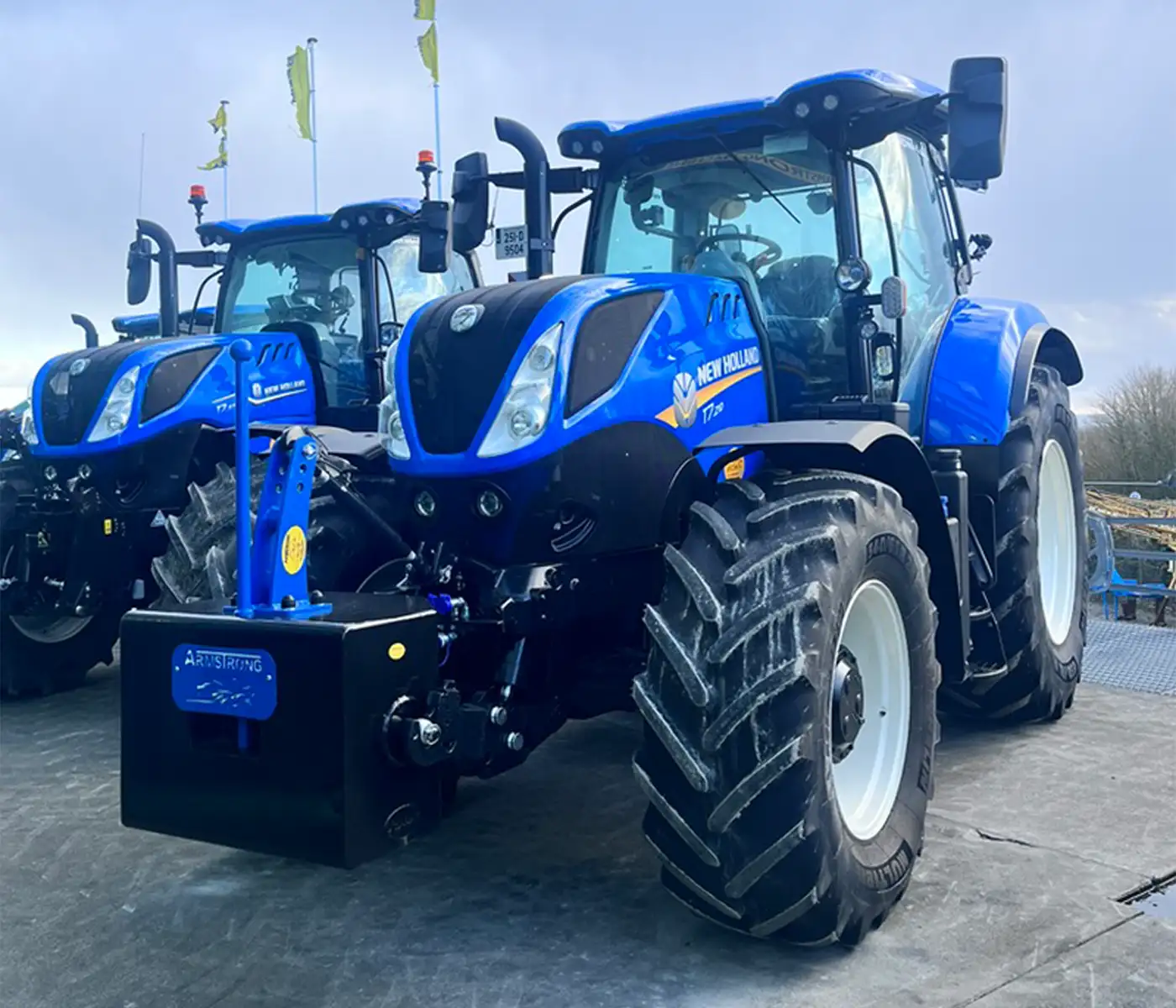 Black and blue weight block attached to a blue New Holland trator beside another Blue New Holland Tractor in a yard