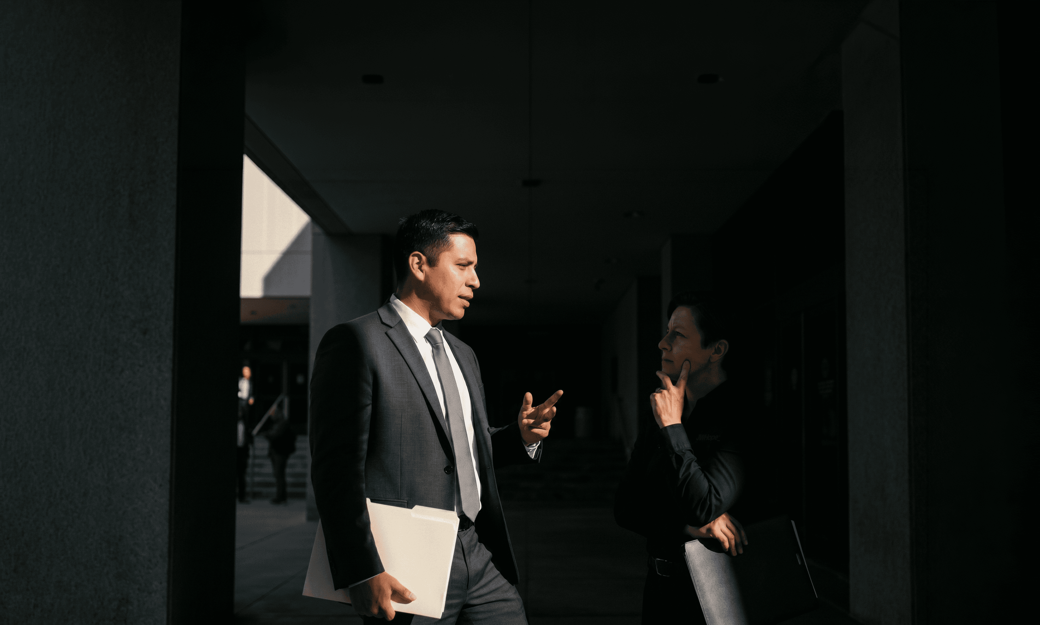 Two individuals in formal business attire engage in conversation in a dim hallway, one holding a document folder, emphasizing a professional setting.