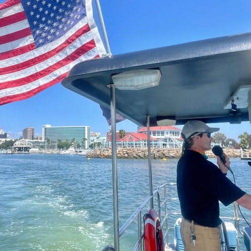 A person on a boat speaks into a microphone, with an American flag flying above. City buildings and a waterfront are in the background.