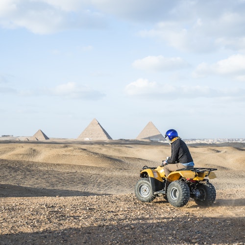 Person riding a yellow ATV on sandy terrain with pyramids visible in the background under a partly cloudy sky.