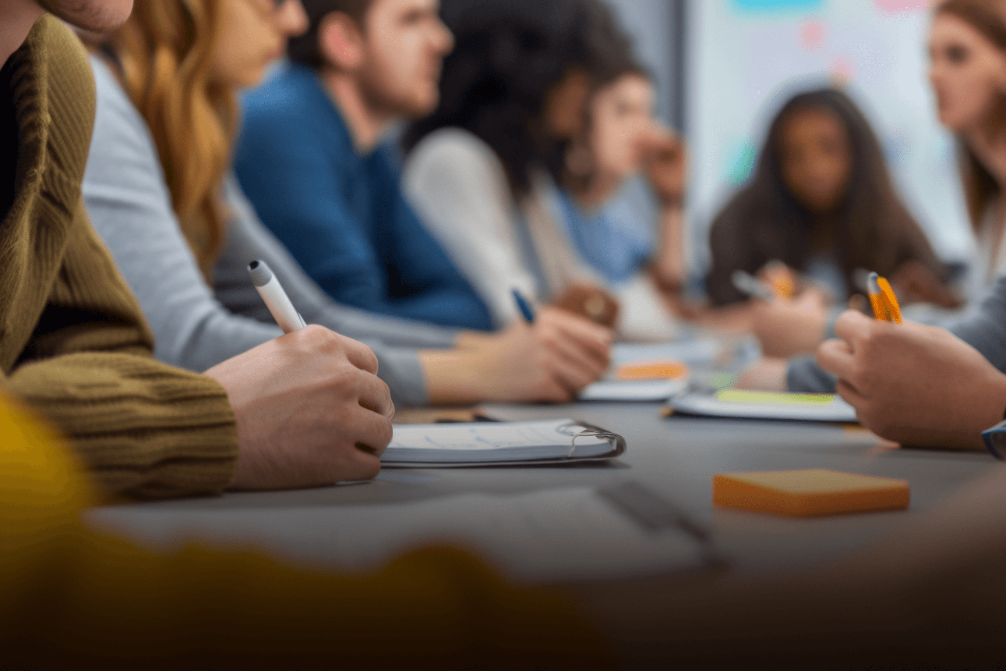 Imagem de um ambiente colaborativo com pessoas reunidas em uma mesa. No foco, mãos segurando canetas e preenchendo pranchetas e papéis, sugerindo um contexto de workshop, brainstorming ou reunião. Ao fundo, outras pessoas desfocadas interagem em um cenário iluminado.