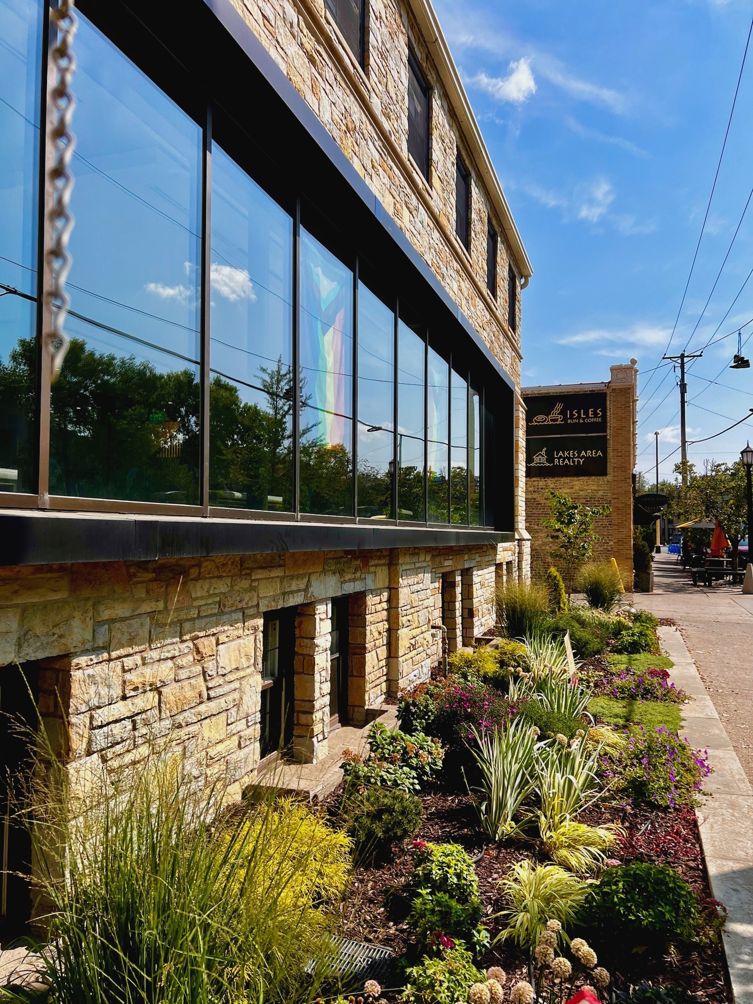 Exterior of Grace Trinity Church with large reflective windows, stone facade, and a well-maintained garden featuring various plants and flowers, with a Pride flag visible through the window