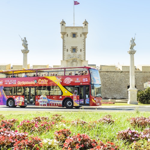 Ein roter Doppeldecker-Tourbus fährt an einem historischen Gebäude mit Statuen und einer Flagge vorbei, umgeben von lebendigen Blumen und Grünflächen.
