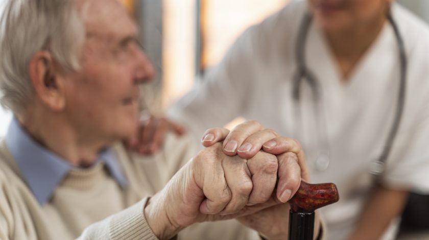 Elderly man holding a cane receives support from a caregiver in a white uniform, symbolizing elderly care and compassion in a healthcare setting.