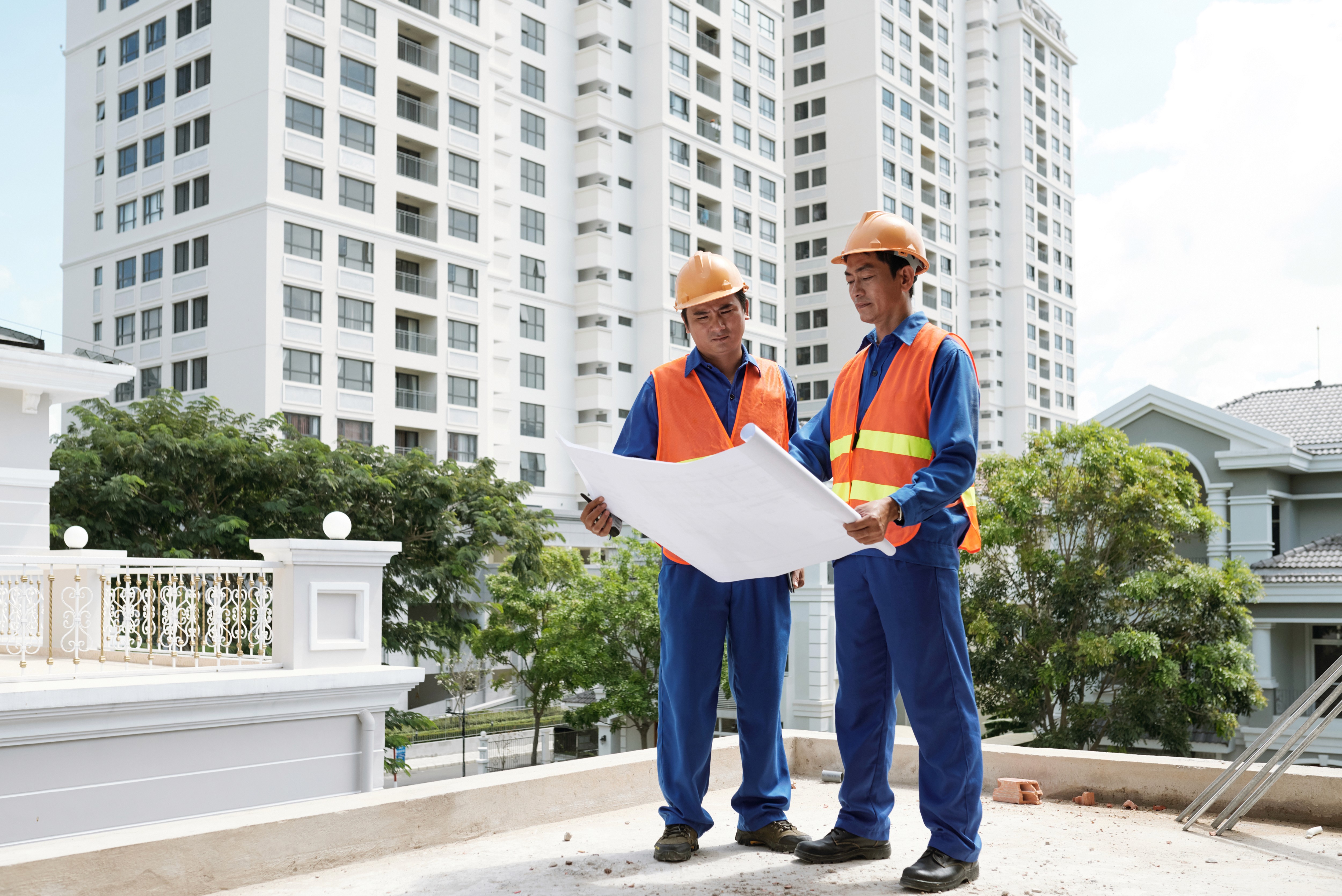 Two construction workers in safety gear review blueprints at a building site. High-rise apartments and trees are visible in the background.