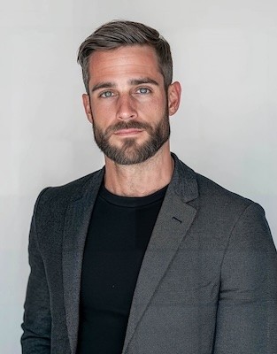 A man with a beard and short hair, wearing a black shirt and gray blazer, posing against a light background.