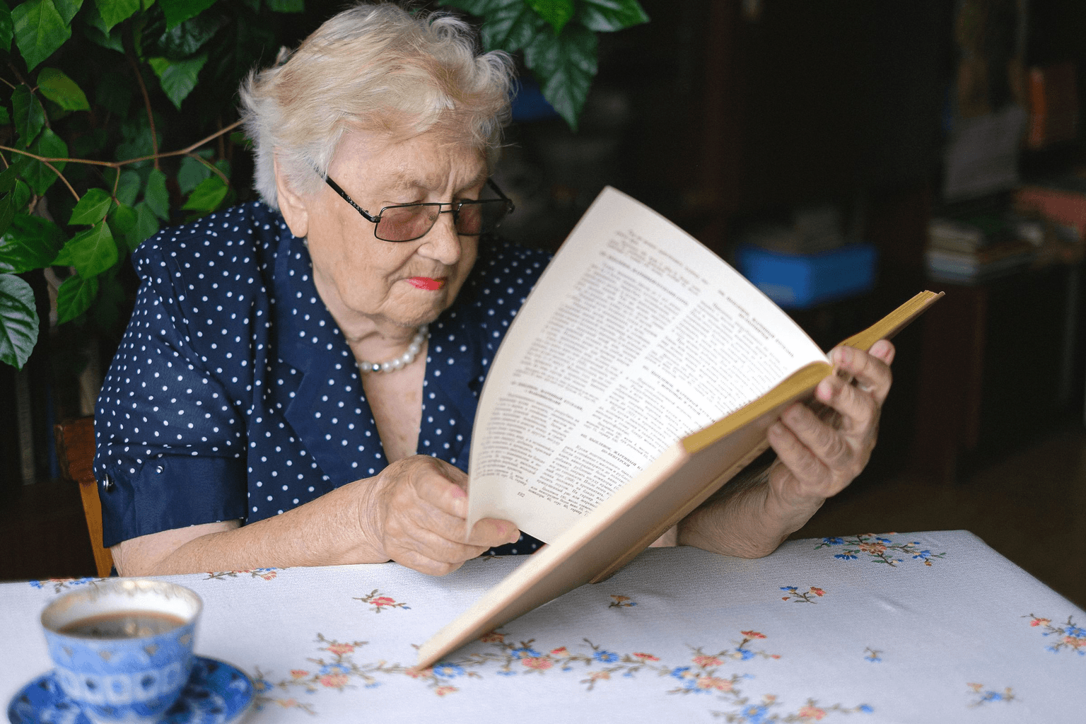 An older woman reading a book at her table with a cup of coffee beside her.