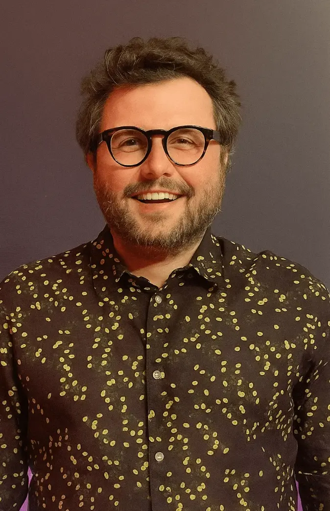Man with glasses and patterned shirt smiling. Co-criador do Tá na História. Thiago Gomide. Fernanda Arouca