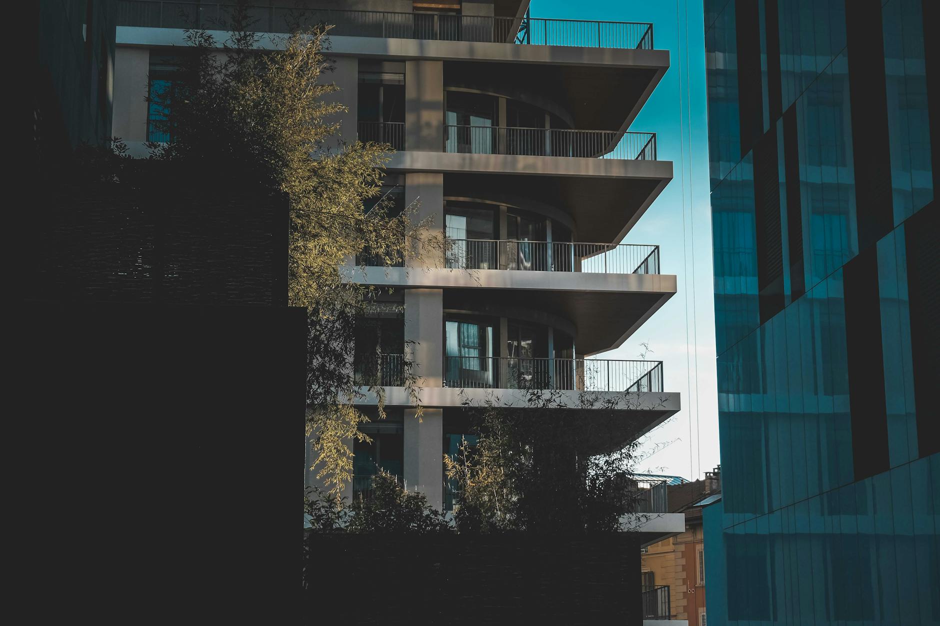 Multi-storey residential apartment block with balconies and trees flanked by dark glass commercial buildings under a blue sky