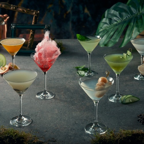 A variety of colorful cocktails in martini glasses on a gray surface with tropical leaves in the background.