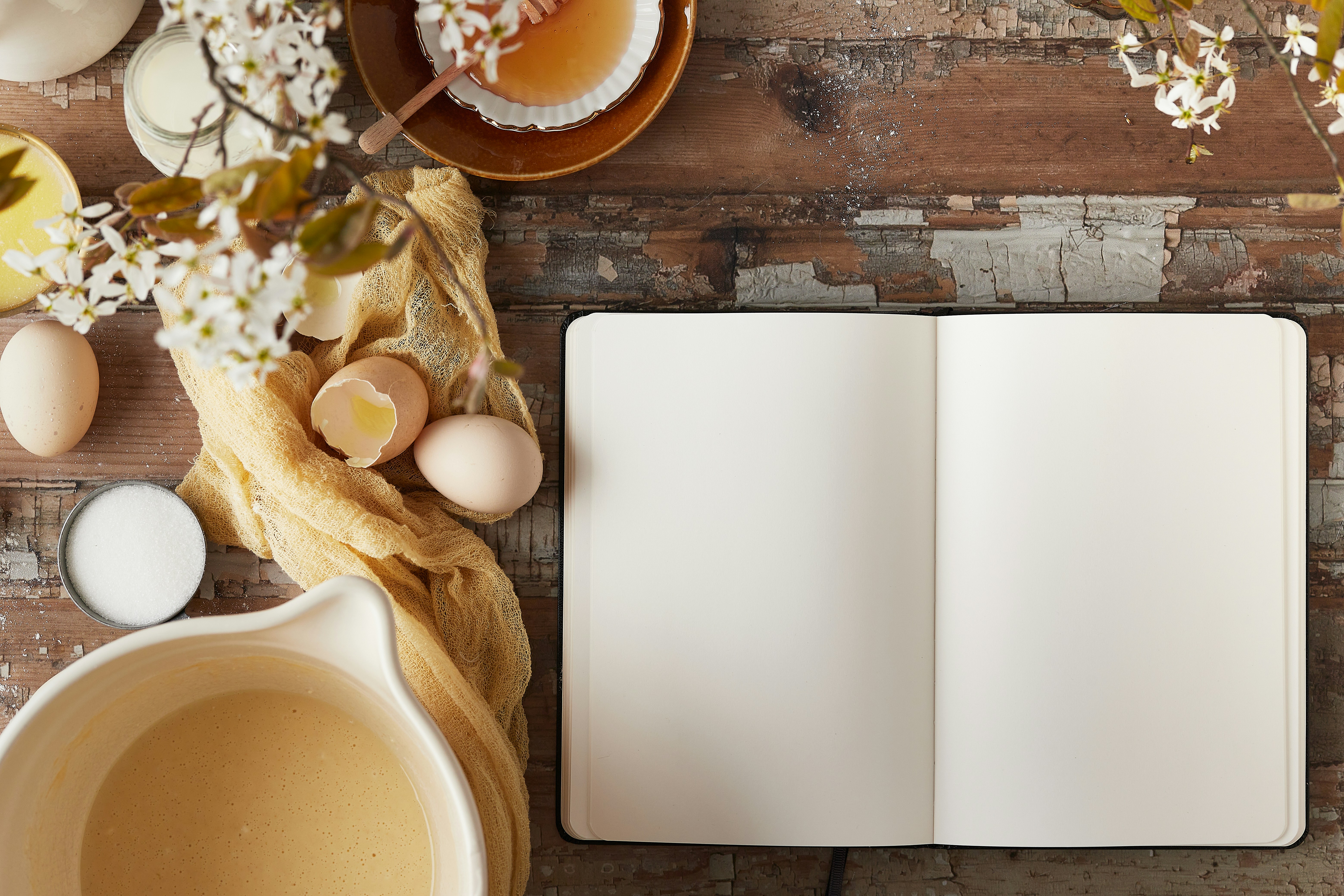 Flat lay of baking ingredients and a blank recipe notebook on a rustic table.