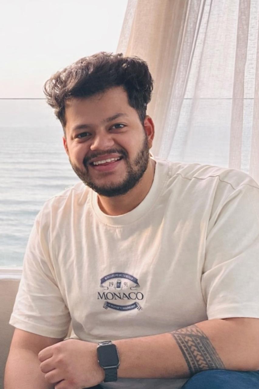 A young woman with long hair smiles while standing against a green backdrop, wearing a casual dark sweater.