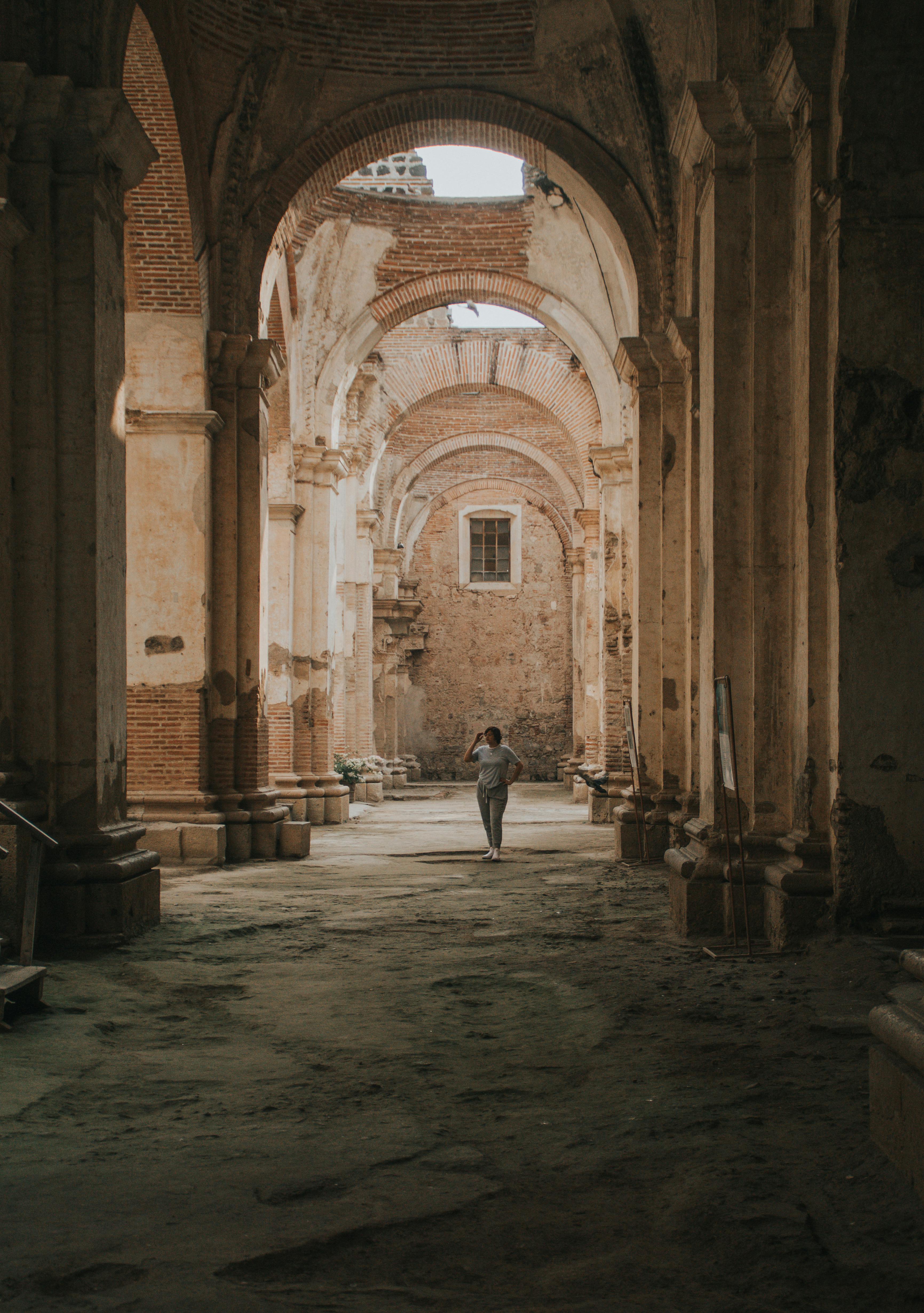 A solitary figure walks through the sunlit ruins of a historic brick and stone cathedral, showcasing its majestic arches and weathered walls.