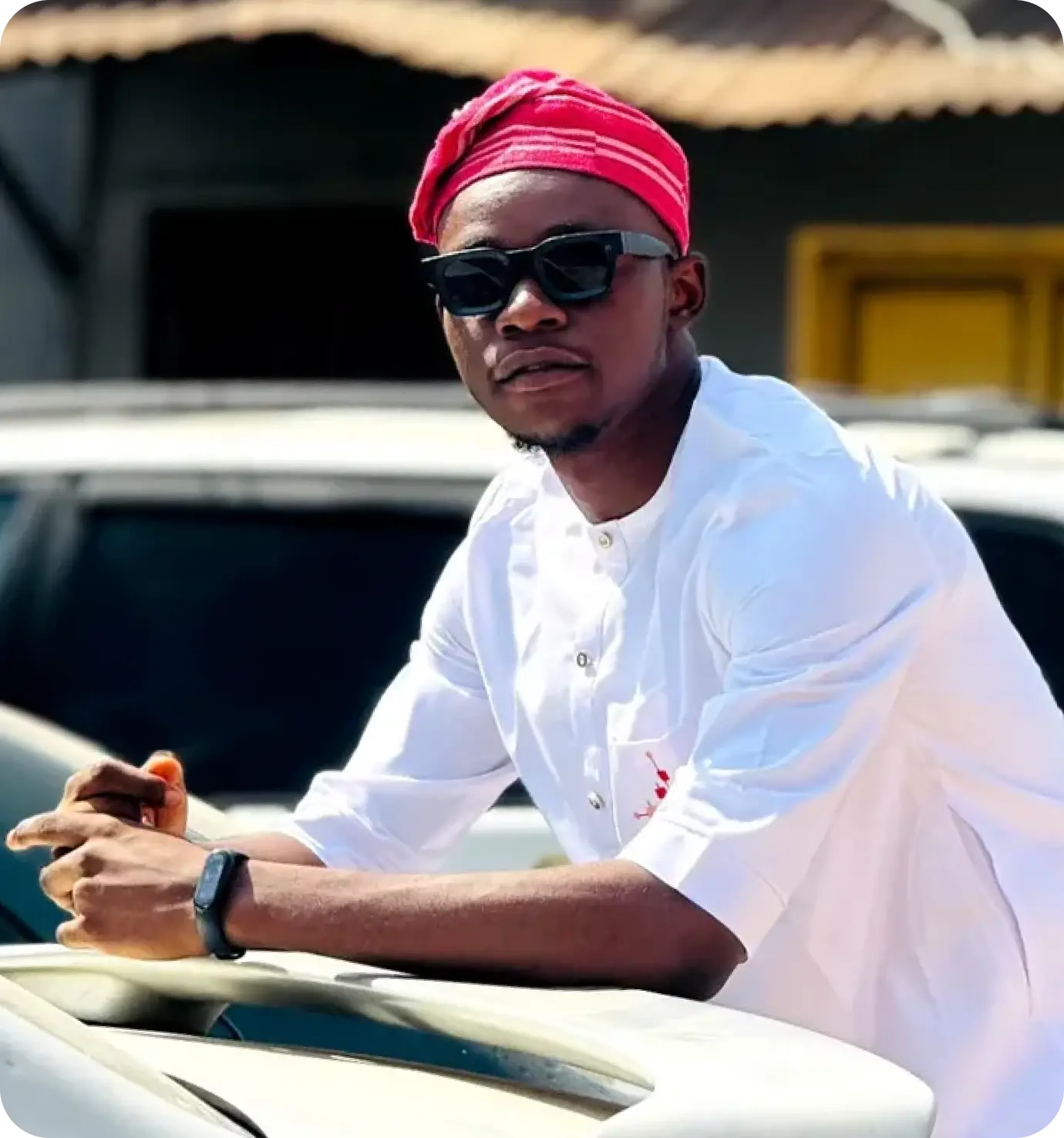 A young man in a plain white traditional apparal and a pink yoruba native cap placed on his head. He is putting on a shade and looks directly at the camera.