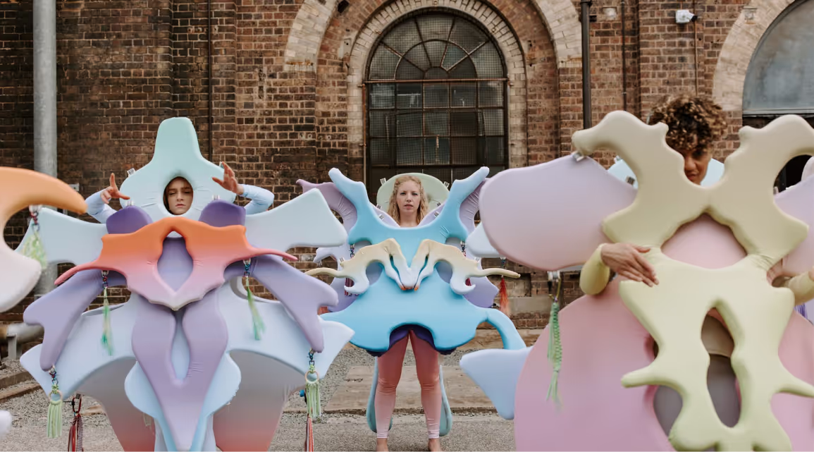 Three women dressed in pastel coloured, symmetrical costume for a performance in front of Carriageworks building.
