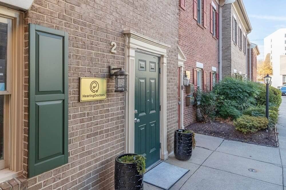 A brick exterior featuring a green door with shutters, a sign, and surrounding greenery on a well-maintained path.
