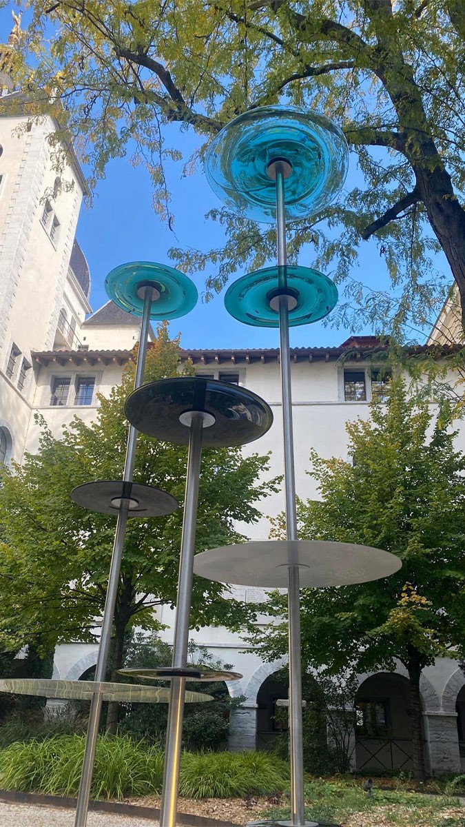 Une installation artistique extérieure composée de plusieurs structures verticales en métal, ressemblant à de fines tiges, surmontées ou agrémentées de disques horizontaux aux couleurs variées, incluant des tons de bleu translucide et de noir opaque. La sculpture est située dans une cour arborée, avec en arrière-plan un bâtiment ancien aux murs blancs doté d'arcades, sous un ciel bleu partiellement visible à travers les feuillages des arbres.