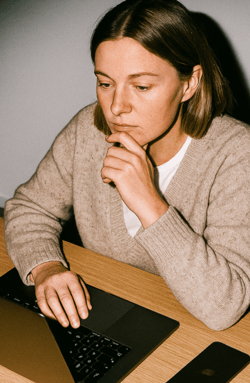 A woman with long brown hair sits at a table, deep in thought with her hand on her chin.