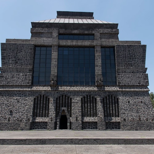 Isang malaking gusali na yari sa bato na may mga parisukat na bintana at pasukang may arko, sa ilalim ng maliwanag na asul na langit.