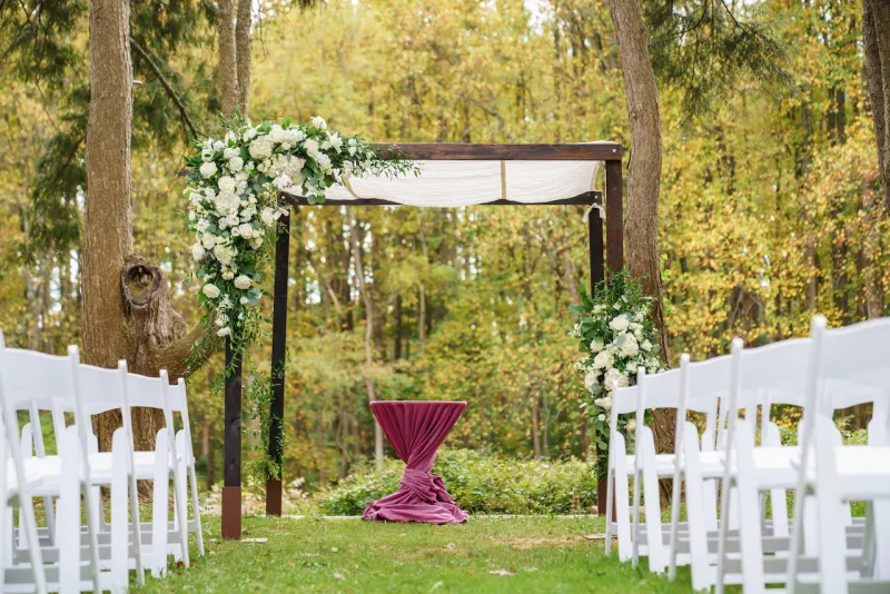 A serene outdoor wedding setup with white chairs arranged on green grass under a canopy of trees.