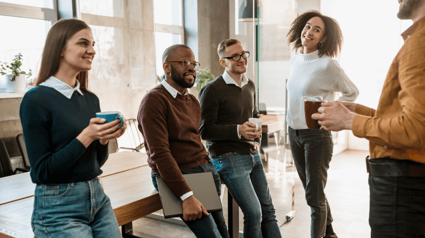 A group of five colleagues stand together in a modern office setting, smiling and holding coffee mugs, suggesting a casual and friendly atmosphere for a team meeting or break.