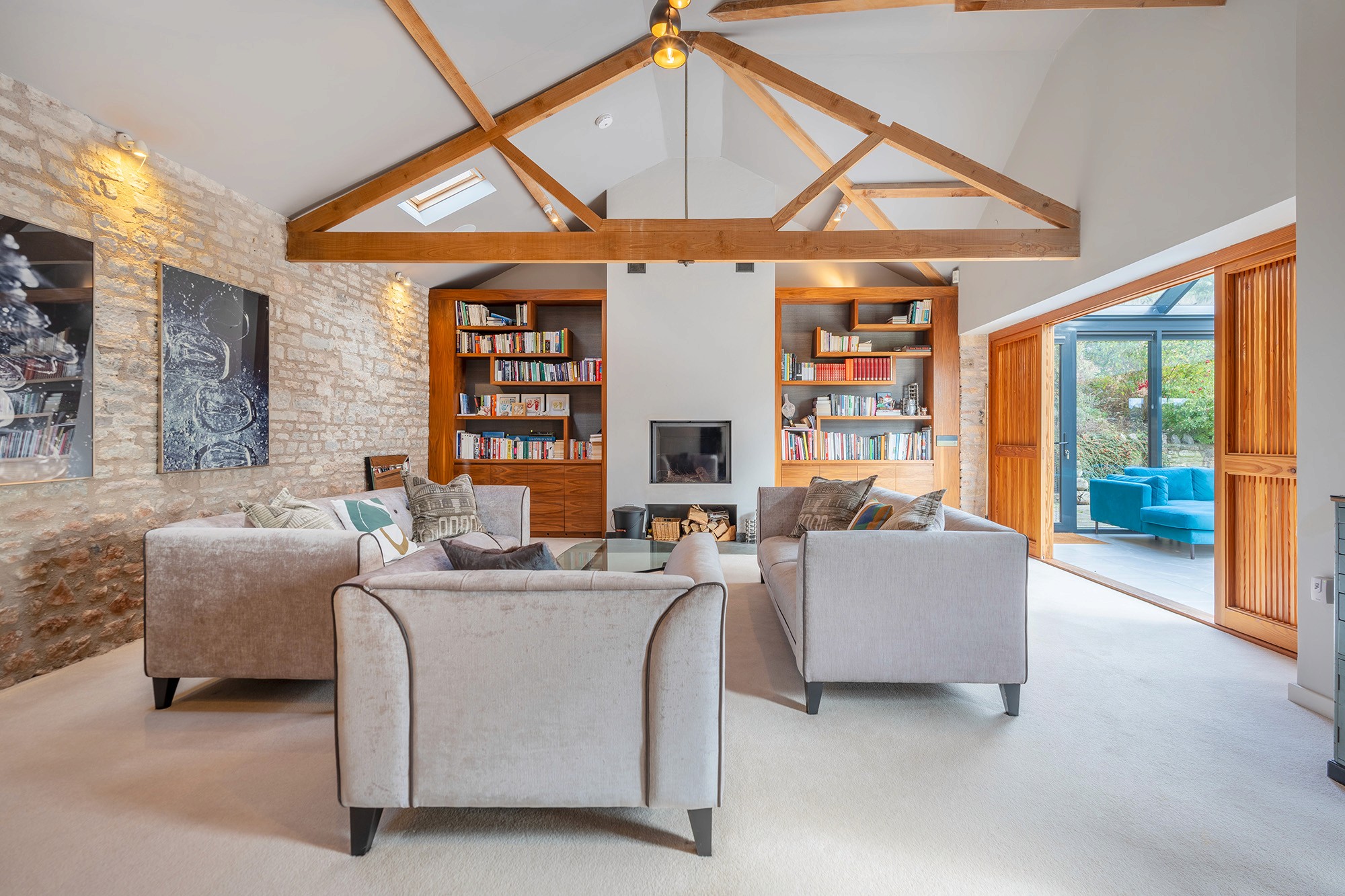 Cozy, high-ceiling living room with exposed wooden ceiling trusses and two pale grey sofas. Features include an exposed stone accent wall, a modern fireplace, and built-in bookshelves flanking the chimney breast. The room opens through wooden slatted doors into the bright sunroom shown in image 2.