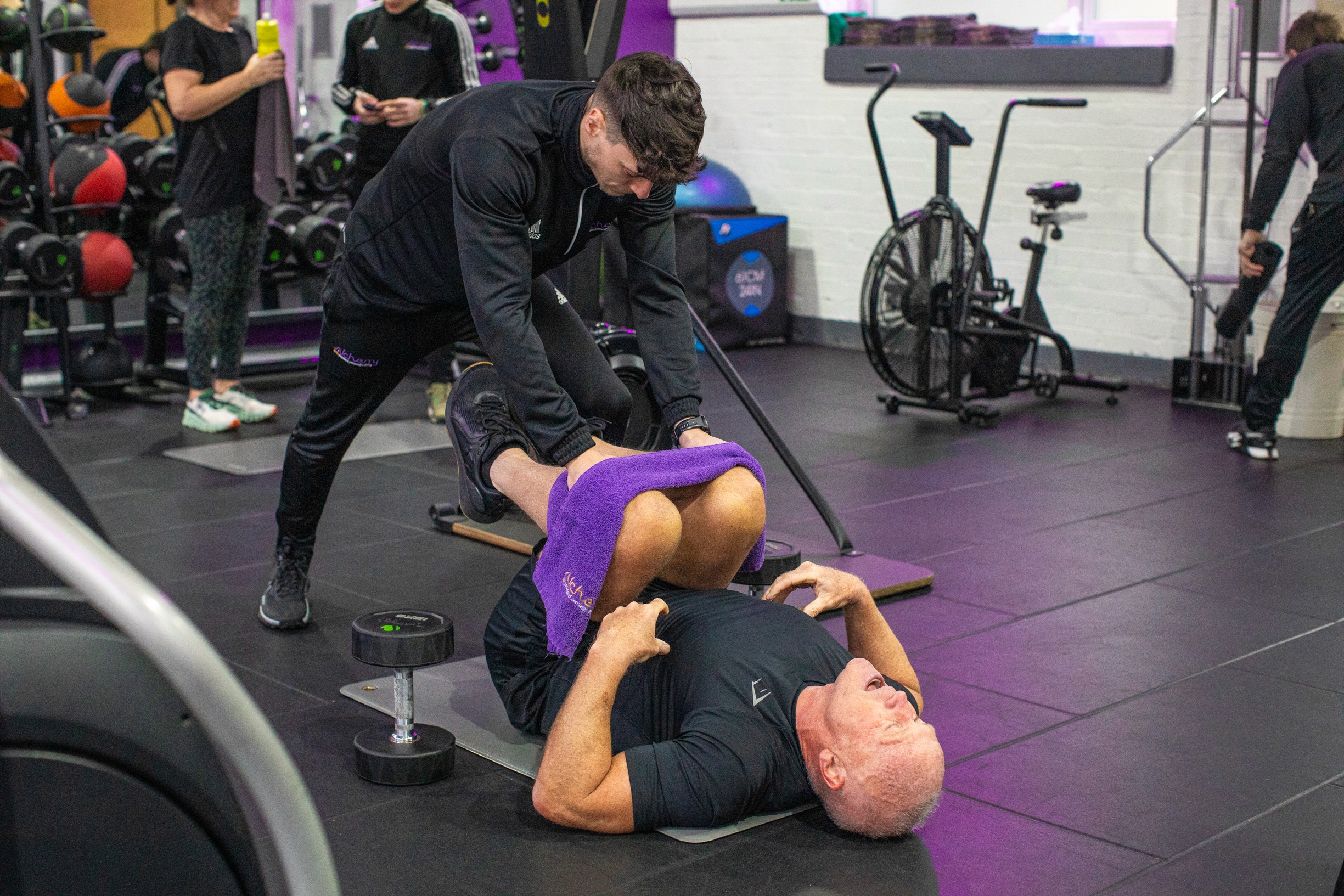 A personal trainer assists an older man in a gym by holding his legs as he does core exercises on the floor using a towel. The atmosphere is focused and supportive.