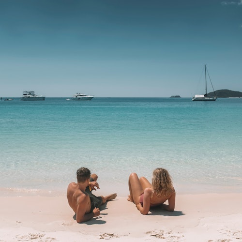 一對夫婦和一個孩子坐在沙灘上,面對著平靜的藍色海洋,遠處有幾艘船停泊。