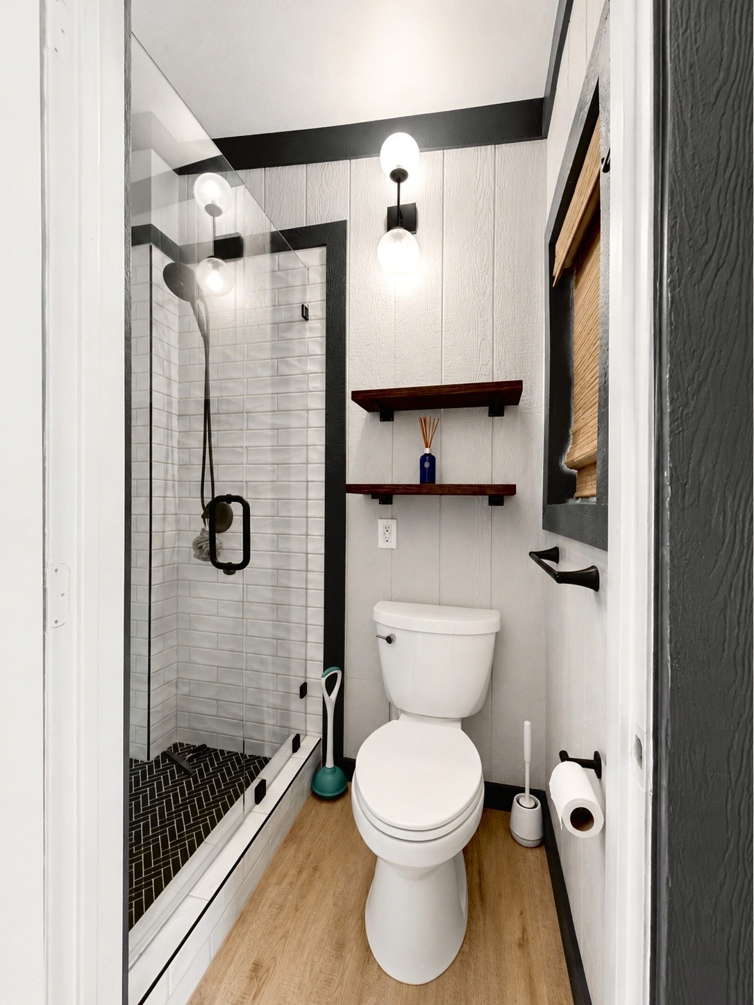 A modern bathroom features a glass-enclosed shower with black fixtures, a white toilet, wooden shelves with a blue vase, and light wood flooring, illuminated by a round bulb wall sconce.