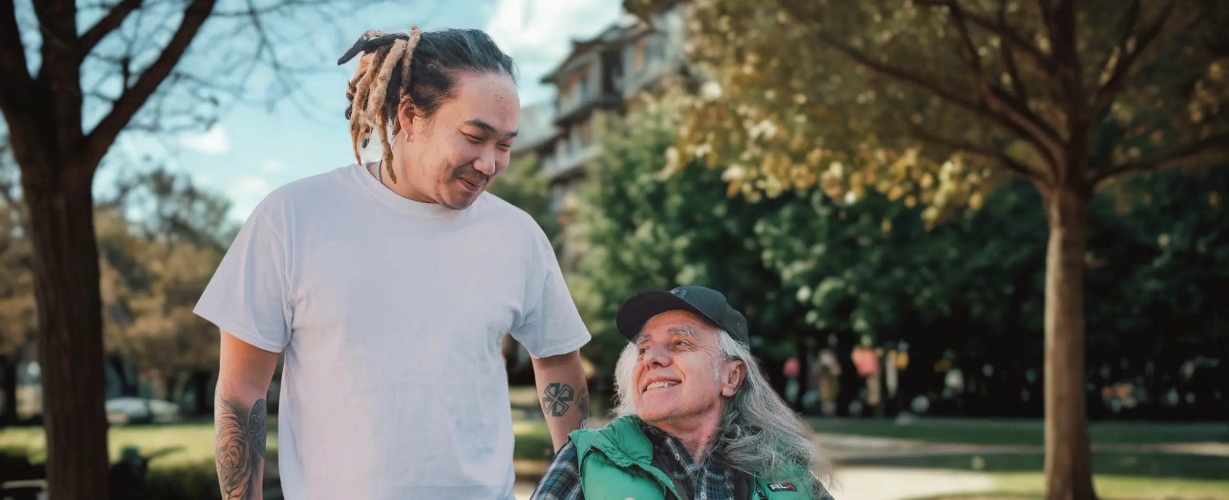 A younger man and an older adult in a wheelchair sharing a lighthearted, laughing moment together outdoors on a sunny day.