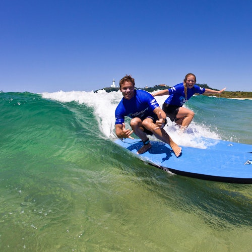 Dalawang tao na nakasuot ng asul na damit ang nagsu-surf sa berdeng alon, na may klarong asul na langit at malayong baybayin sa likuran.