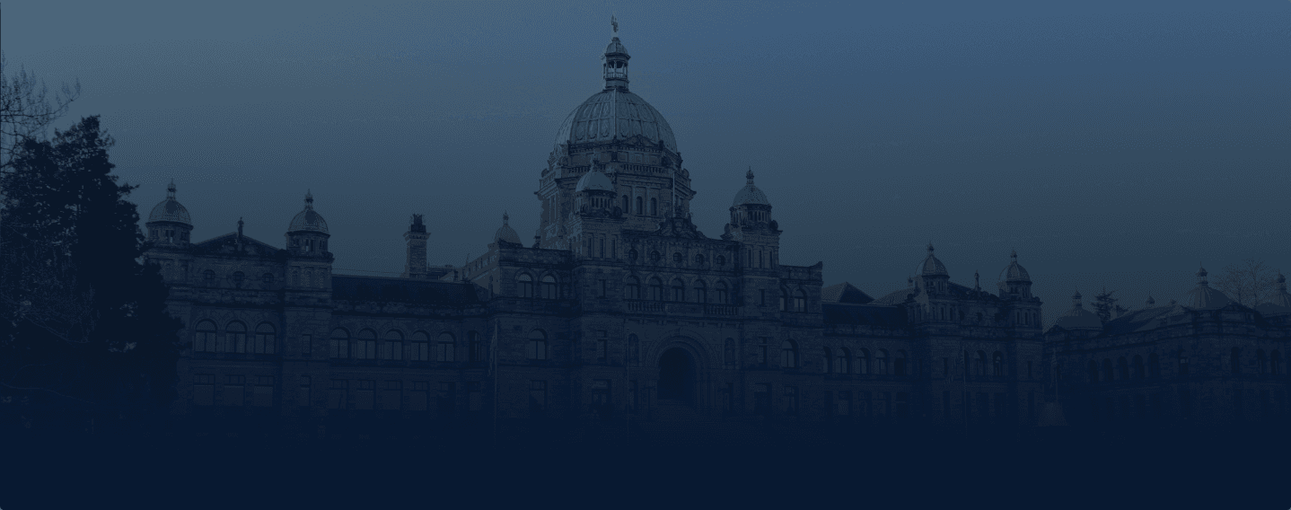 The image shows a majestic Victoria, BC parliament  building with a large central dome and several smaller towers, silhouetted against a darkening sky, possibly during twilight.
