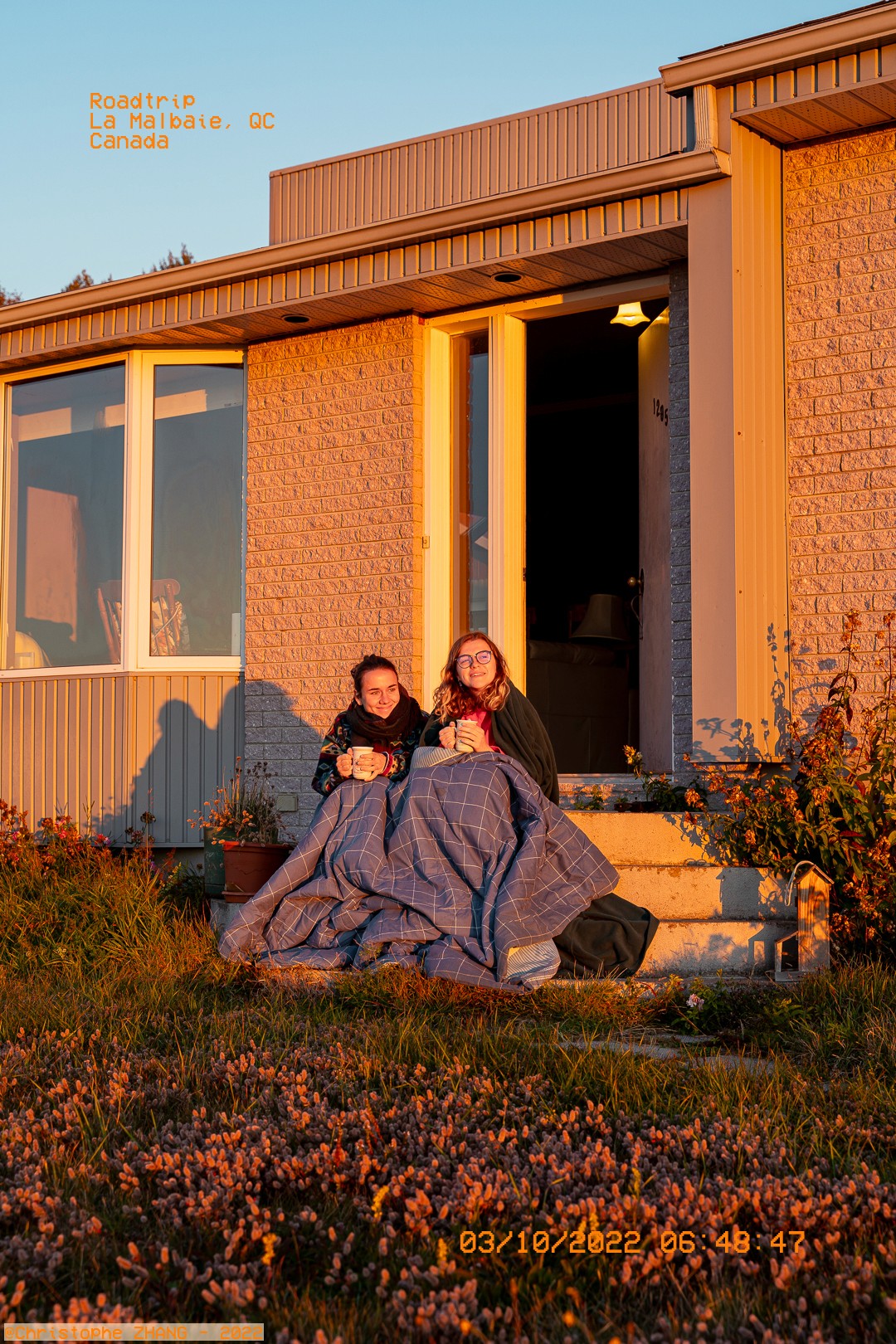 Deux femmes profitant du lever du soleil, couvertes de couvertures sur le perron d’une maison à La Malbaie